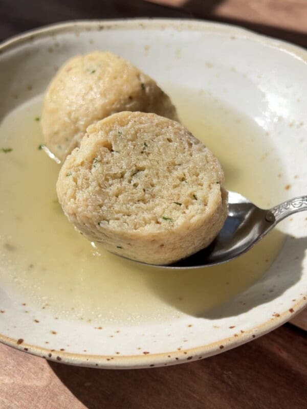 A close-up of a bowl with clear broth and a sliced bread dumpling, one half resting on a spoon. The dumpling appears soft with visible herbs, and sunlight highlights the textures.