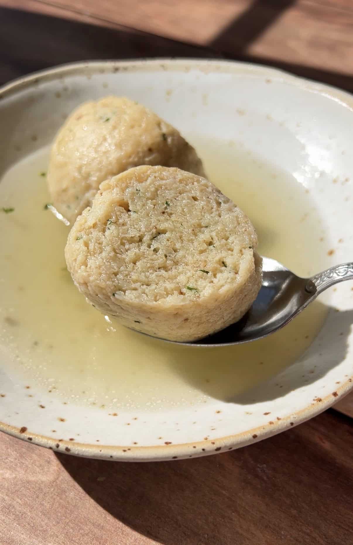 A close-up of a bowl with clear broth and a sliced bread dumpling, one half resting on a spoon. The dumpling appears soft with visible herbs, and sunlight highlights the textures. - 4