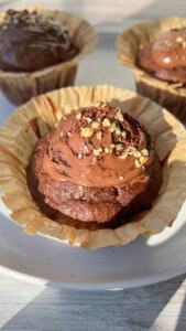 Close-up of a chocolate cupcake in a paper liner, topped with chocolate frosting and sprinkled with chopped nuts. Two more decorated cupcakes are visible in the background on a white plate.