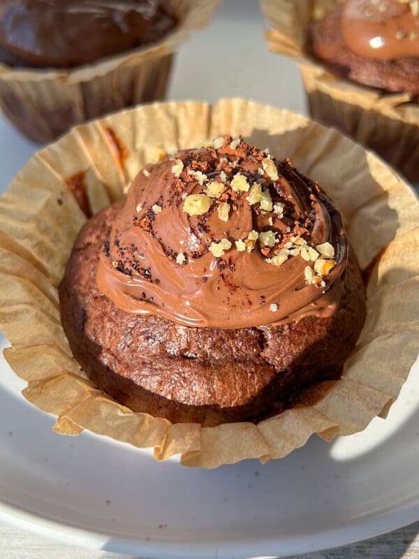 Close-up of a chocolate cupcake in a paper liner, topped with chocolate frosting and sprinkled with chopped nuts. Two more decorated cupcakes are visible in the background on a white plate.