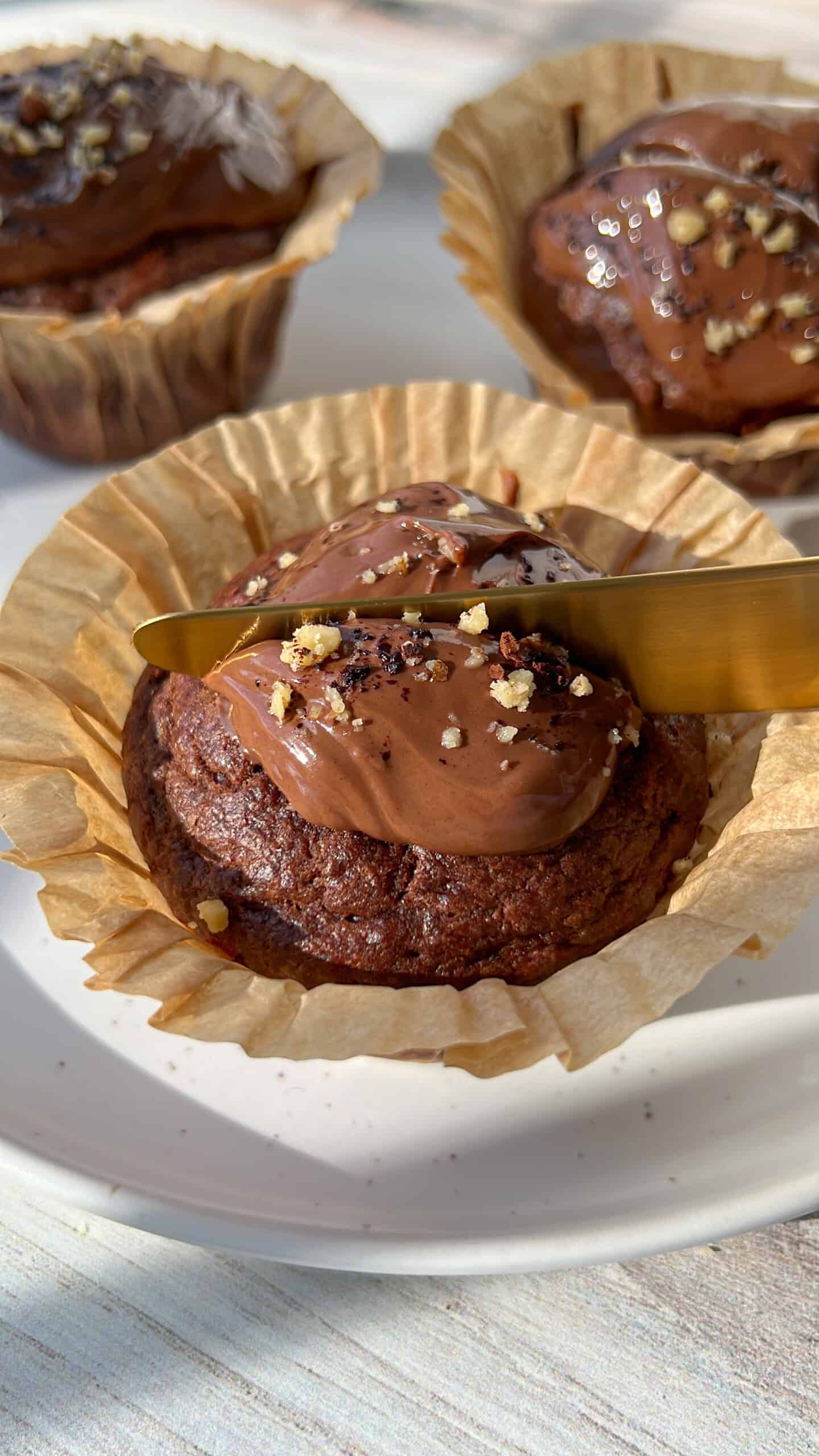 A close-up of a chocolate muffin in a paper liner being spread with chocolate frosting using a gold knife. Two more frosted muffins are visible in the background on a white plate.