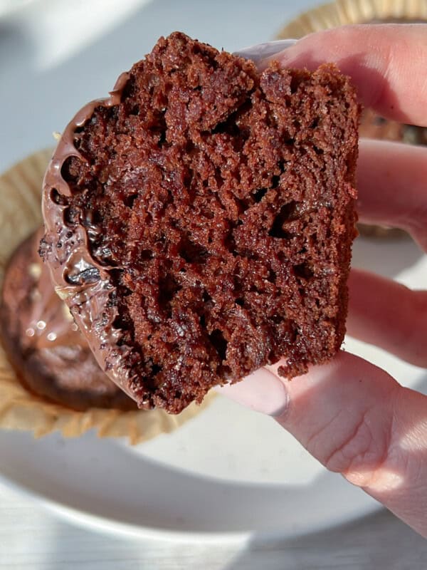 A hand holds a cut chocolate cupcake, showing its moist, rich, and fluffy interior. The cupcake is topped with a layer of melted chocolate icing. In the background, more cupcakes sit on a white plate.