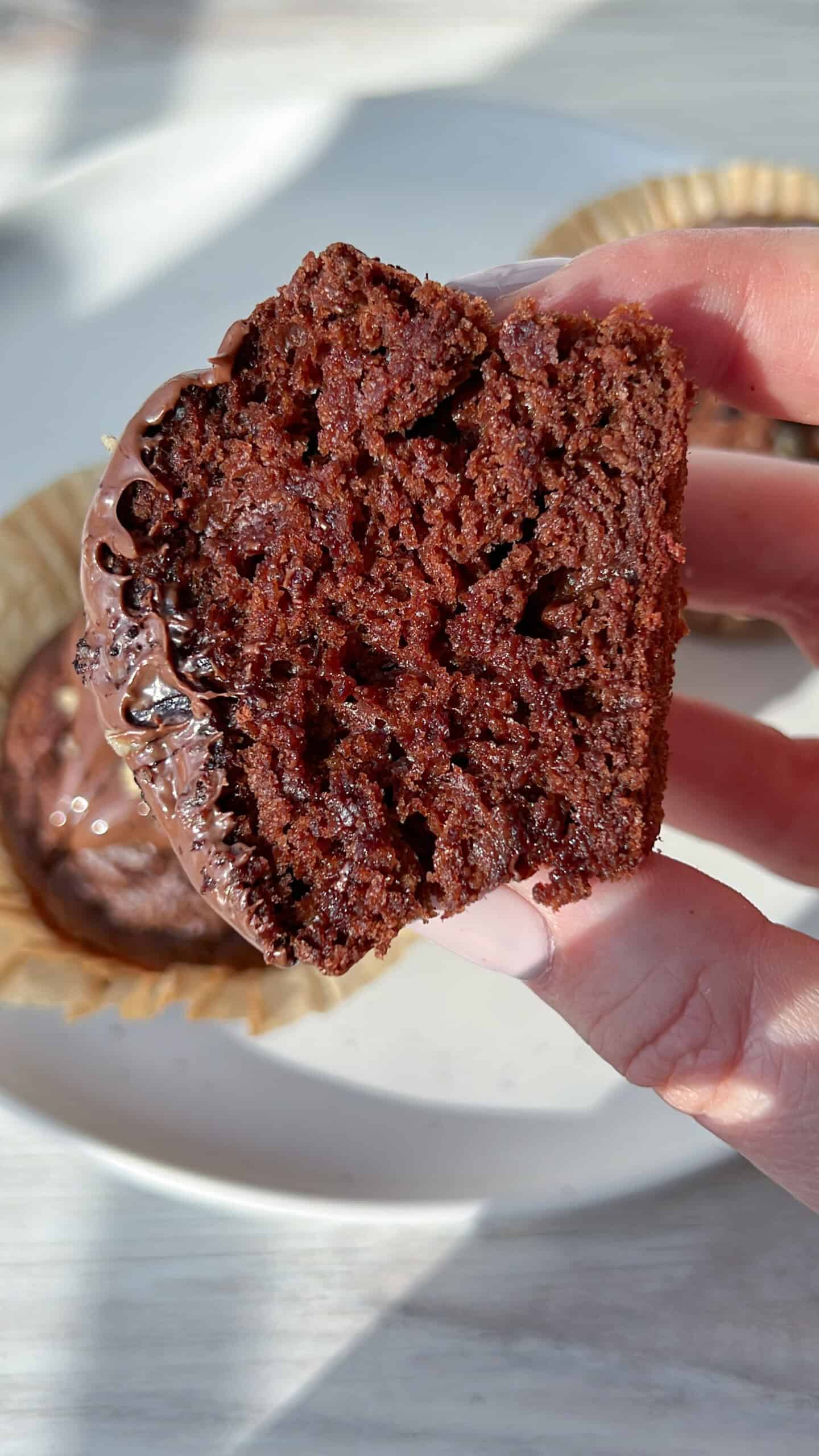 A hand holds a cut chocolate cupcake, showing its moist, rich, and fluffy interior. The cupcake is topped with a layer of melted chocolate icing. In the background, more cupcakes sit on a white plate.