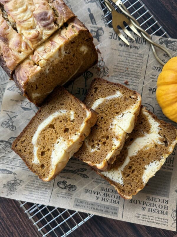 Sliced pumpkin bread with creamy swirls, displayed on vintage-style paper atop a cooling rack, with a small pumpkin and serving utensils nearby.