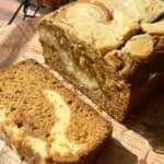 A sliced loaf of pumpkin bread with a cream cheese swirl sits on brown parchment paper. Sunlight highlights the moist texture and marbled swirl. A small decorative pumpkin is in the background.