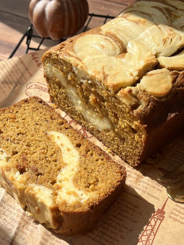 A sliced loaf of pumpkin bread with a cream cheese swirl sits on brown parchment paper. Sunlight highlights the moist texture and marbled swirl. A small decorative pumpkin is in the background.