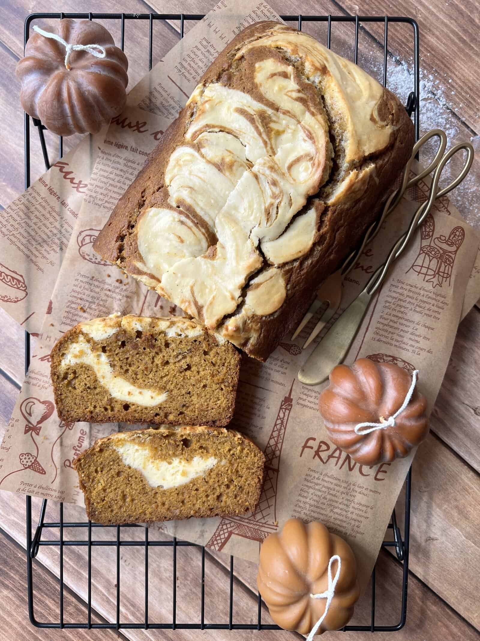 A loaf of pumpkin bread with cream cheese swirls, two slices cut, on a cooling rack with decorative paper, surrounded by three small faux pumpkins and two gold forks. - 4