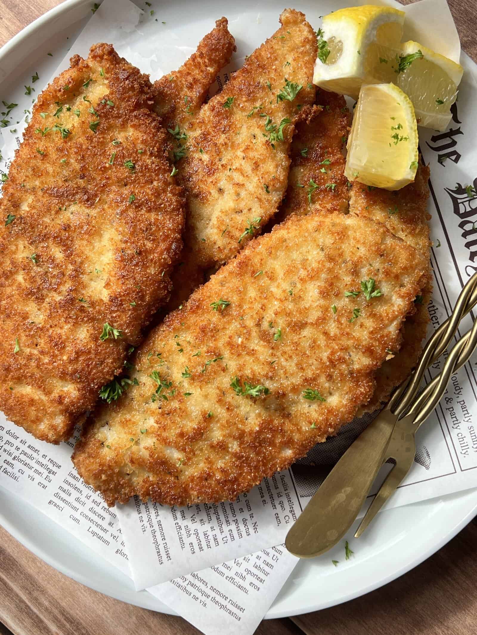 Three golden-brown breaded schnitzels on a white plate, garnished with chopped parsley and served with lemon wedges. A fork and knife rest on the plate, which is lined with printed paper. - 3