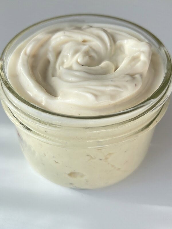 A small glass jar filled with creamy, white whipped spread, possibly mayonnaise or a dip, with a smooth, swirled texture on top, sitting on a white surface.