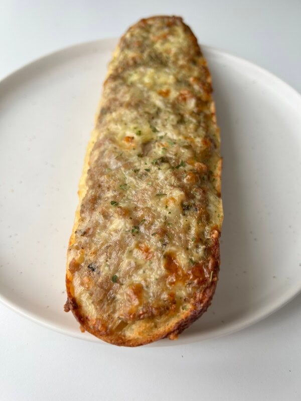 A toasted, cheesy, and herb-topped baguette half sits on a round white plate against a light background.