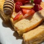 A close-up of sliced cheesecake topped with fresh strawberries, raspberries, and lemon slices, drizzled with honey from a wooden honey dipper, served on a white plate in sunlight.