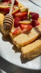A close-up of sliced cheesecake topped with fresh strawberries, raspberries, and lemon slices, drizzled with honey from a wooden honey dipper, served on a white plate in sunlight.
