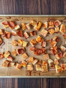 A baking sheet covered with parchment paper holds many small, square pizza croutons with various toppings, perfect for making a flavorful Pizza Crouton Salad, all spread out over a wooden surface.