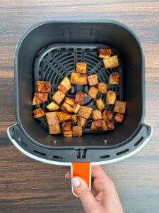 A hand holds an air fryer basket containing crispy, golden-brown cubes of tofu, perfect as a protein-packed topping for a Pizza Crouton Salad, displayed on a wooden surface.