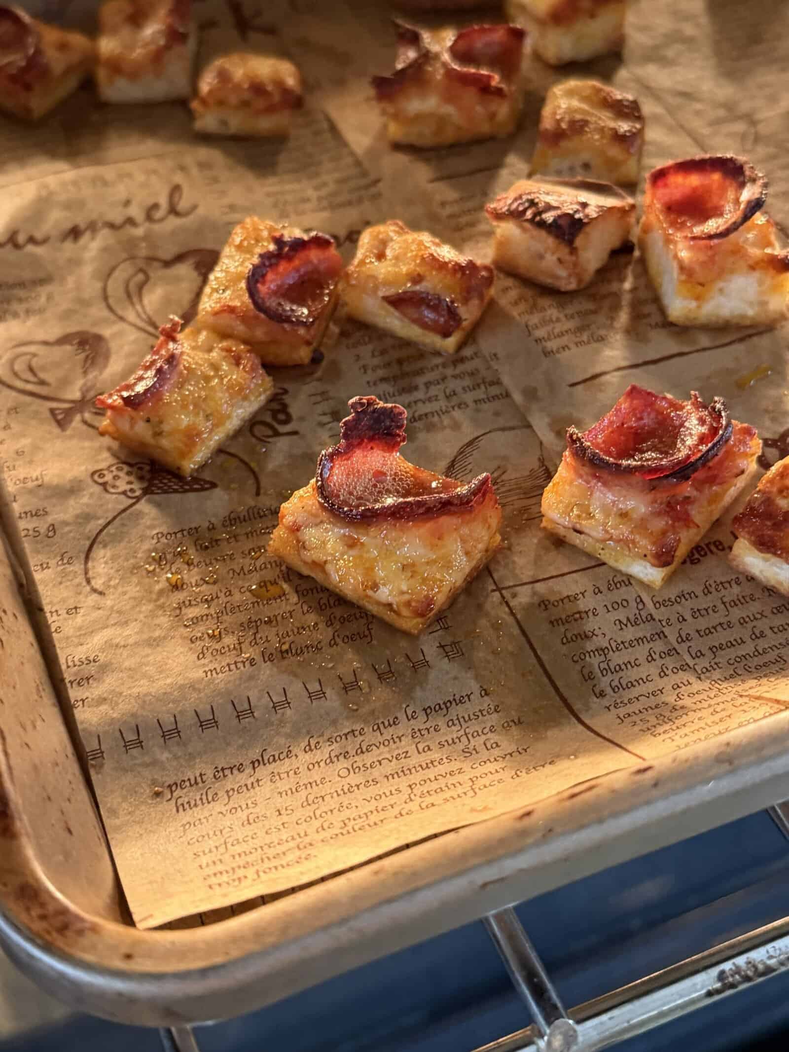 Small, square pieces of toasted bread topped with melted cheese and crispy bacon are arranged on a parchment-lined baking sheet inside an oven—perfect for adding crunch to a Pizza Crouton Salad. The parchment paper is printed with French text and illustrations.