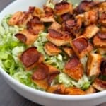 A close-up of a Pizza Crouton Salad in a white bowl, topped with crispy, roasted bread cubes and thin slices of pepperoni. The green salad underneath appears to be chopped lettuce. Two gold-colored utensils rest in the bowl.