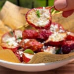 A hand holds up a halved stuffed cherry pepper over a bowl lined with paper, filled with more stuffed peppers topped with crumbled cheese and herbs, outdoors in natural sunlight.