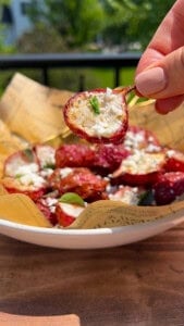 A hand holds up a halved stuffed cherry pepper over a bowl lined with paper, filled with more stuffed peppers topped with crumbled cheese and herbs, outdoors in natural sunlight.