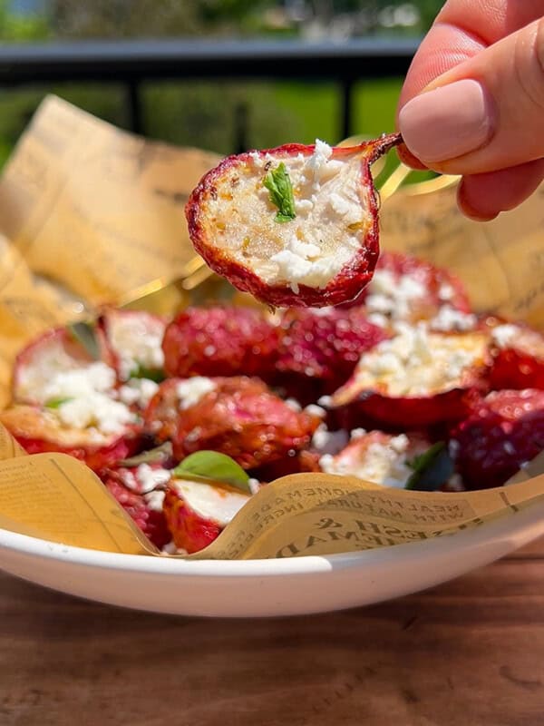 A hand holds up a halved stuffed cherry pepper over a bowl lined with paper, filled with more stuffed peppers topped with crumbled cheese and herbs, outdoors in natural sunlight. - 77
