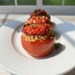 Three roasted tomatoes stuffed with rice and herbs are arranged on a white plate, set by a sunlit window with a view of a green garden in the background.