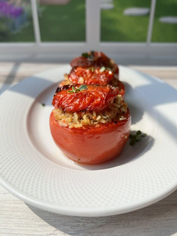 Three roasted tomatoes stuffed with rice and herbs are arranged on a white plate, set by a sunlit window with a view of a green garden in the background.