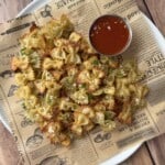 A plate of crispy bowtie pasta chips, reminiscent of Pizza Crouton Salad, garnished with herbs and served on decorative parchment paper with a small cup of red dipping sauce on the side, placed on a wooden table.