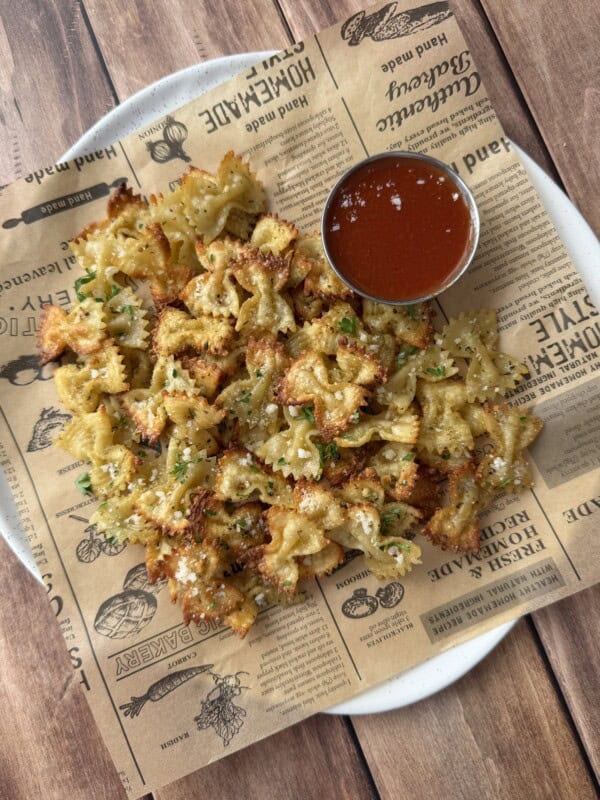 A plate of crispy bowtie pasta chips, reminiscent of Pizza Crouton Salad, garnished with herbs and served on decorative parchment paper with a small cup of red dipping sauce on the side, placed on a wooden table.
