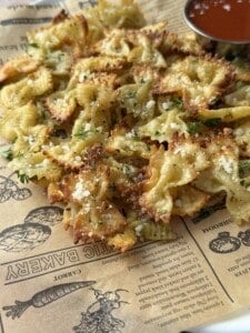 A plate of crispy baked bowtie pasta, like pizza croutons, sprinkled with grated cheese and herbs, served on parchment paper with a small metal cup of red dipping sauce. Perfect as a crunchy topping for your favorite Pizza Crouton Salad.