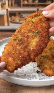 A hand holding a golden-brown, crispy breaded cutlet garnished with chopped parsley over a plate lined with newspaper-style parchment paper. More cutlets are visible on the plate in the background.