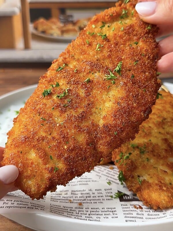 A hand holding a golden-brown, crispy breaded cutlet garnished with chopped parsley over a plate lined with newspaper-style parchment paper. More cutlets are visible on the plate in the background.
