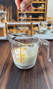 A hand sprinkles salt into a glass measuring cup containing a white liquid, likely cream or milk, on a wooden table with a blurred background of baked goods and what could be ingredients for Pizza Crouton Salad.