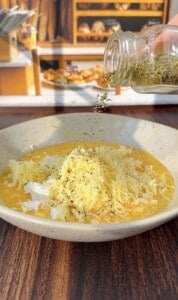 A hand sprinkles dried herbs from a jar onto a bowl of beaten eggs topped with shredded cheese, with a bakery display blurred in the background.