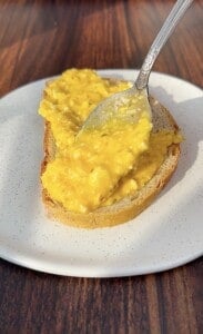 A spoon spreads creamy scrambled eggs onto a slice of bread on a white plate, set on a wooden surface.