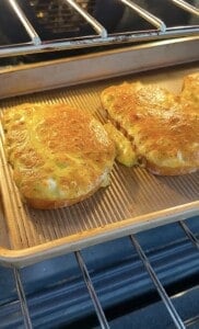 Two golden-brown, seasoned pieces of bread or toast are baking on a ridged baking sheet inside an oven, with a melted and crispy surface.