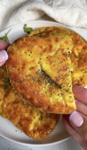 A hand holding a golden, herb-topped cheesy bread with a crispy crust above a plate with more pieces of the same bread. A light cloth is visible in the background.