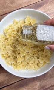 A hand sprinkles dried oregano from a Simply Organic jar over a bowl of cooked farfalle pasta on a wooden table.
