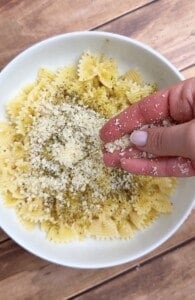 A hand sprinkles grated cheese over a bowl of farfalle pasta topped with herbs, on a wooden surface.