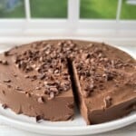 A round chocolate cake with a slice missing, topped with chocolate shavings, sits on a white plate in front of a window with a green garden view in the background.