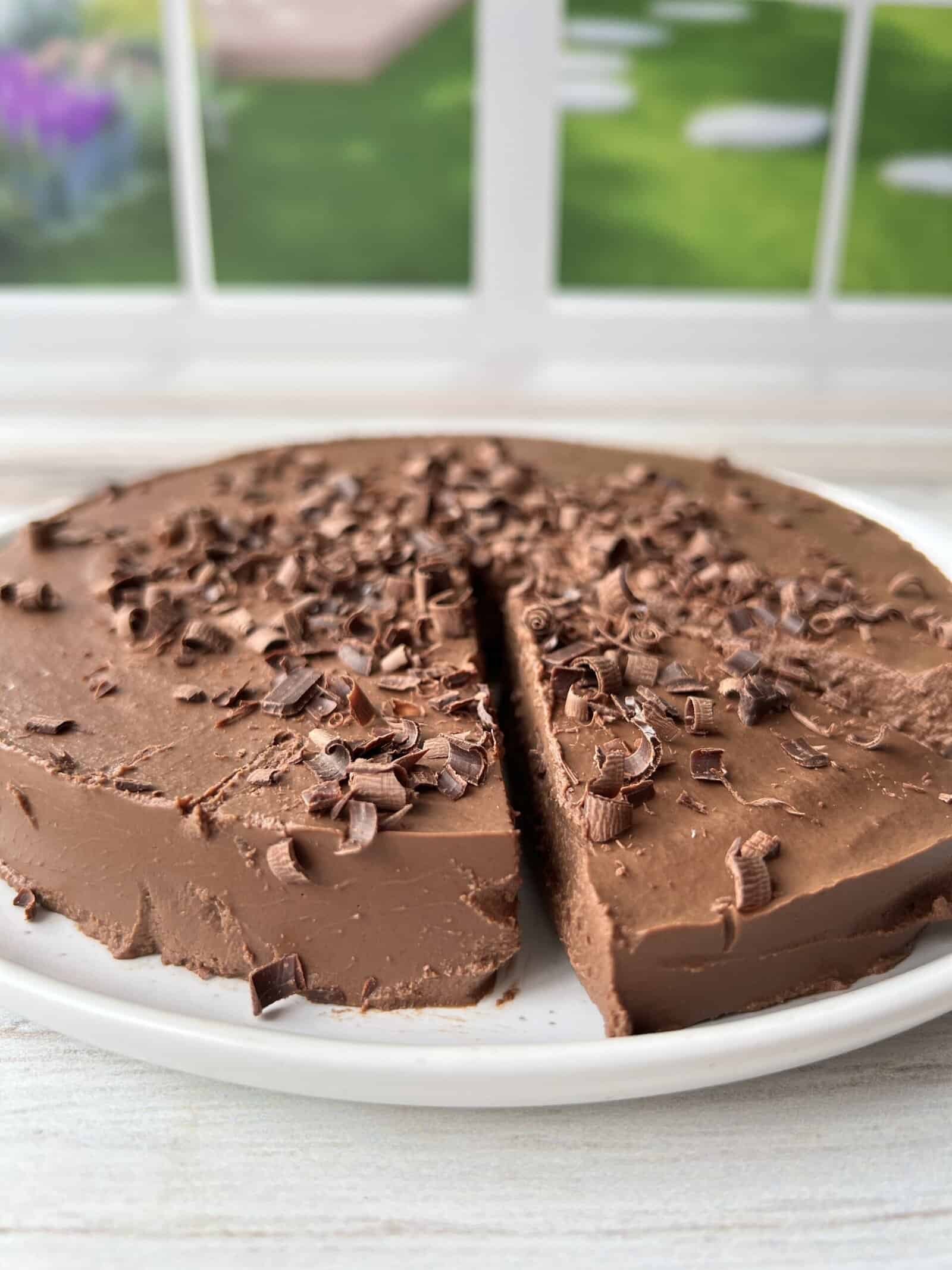 A round chocolate cake with a slice missing, topped with chocolate shavings, sits on a white plate in front of a window with a green garden view in the background.