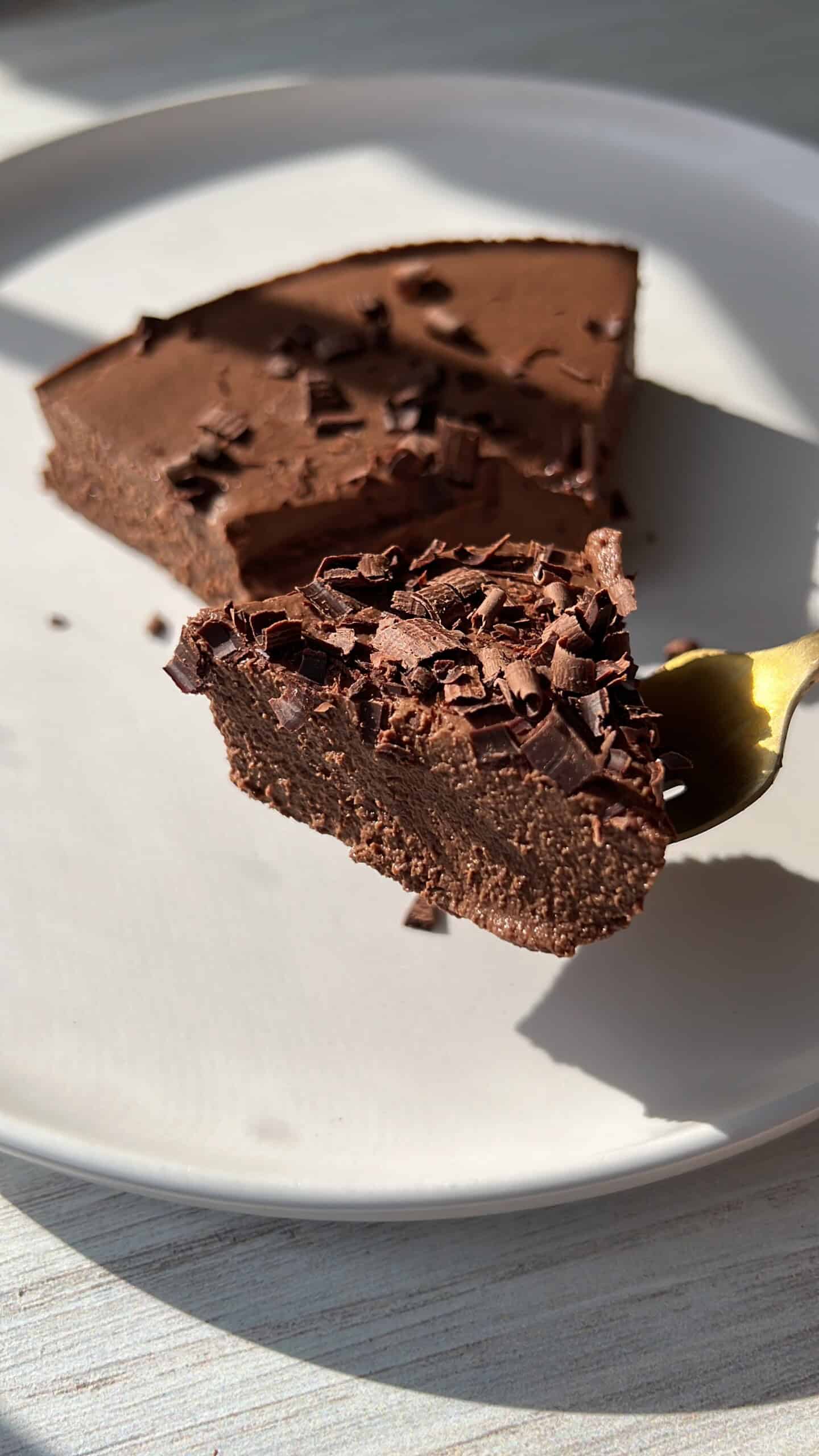 A close-up of a creamy chocolate dessert topped with chocolate shavings, with a fork holding a bite-sized piece above a white plate in natural sunlight.
