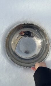 A hand holds a metal bowl partially embedded in snow, with a reflection of a person and buildings on its shiny surface. The scene is bright, set against a soft white snowy background—perfect for making viral snow ice cream.