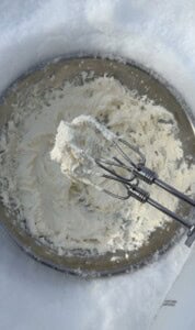 A close-up of an electric mixer with beaters covered in Viral Snow Ice Cream, held above a metal mixing bowl filled with the same creamy mixture.