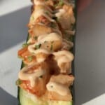 A close-up of a zucchini boat filled with a shrimp mixture, topped with creamy sauce and chopped herbs, served on a white plate with a hand holding one end.