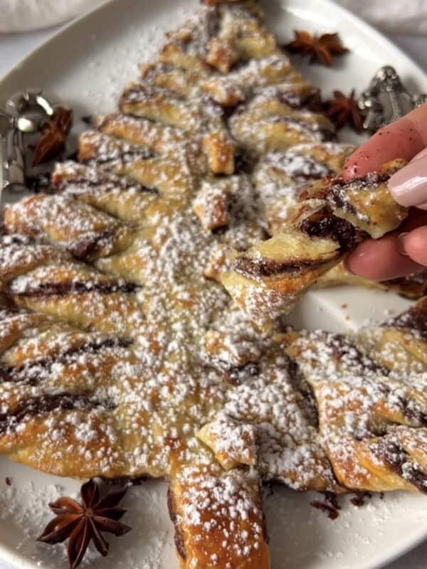 A Christmas tree-shaped pastry dusted with powdered sugar, topped with chocolate spread, is served on a white plate. A hand is pulling off a twisted branch piece. Star anise garnishes the plate.