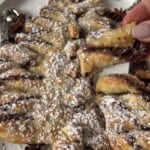 A festive pastry shaped like a Christmas tree, filled with chocolate and dusted with powdered sugar, sits on a plate. A hand holds a piece of the pastry. Star anise decorations are placed around the plate.