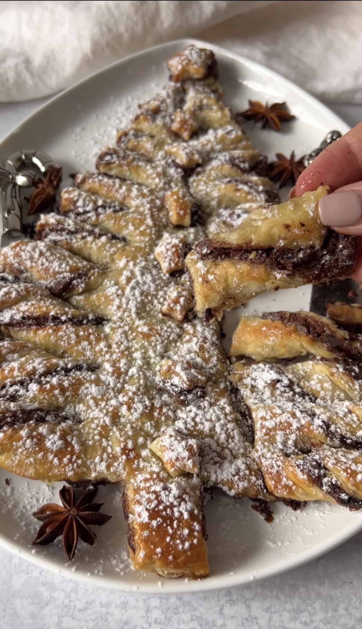 A festive pastry shaped like a Christmas tree, filled with chocolate and dusted with powdered sugar, sits on a plate. A hand holds a piece of the pastry. Star anise decorations are placed around the plate.