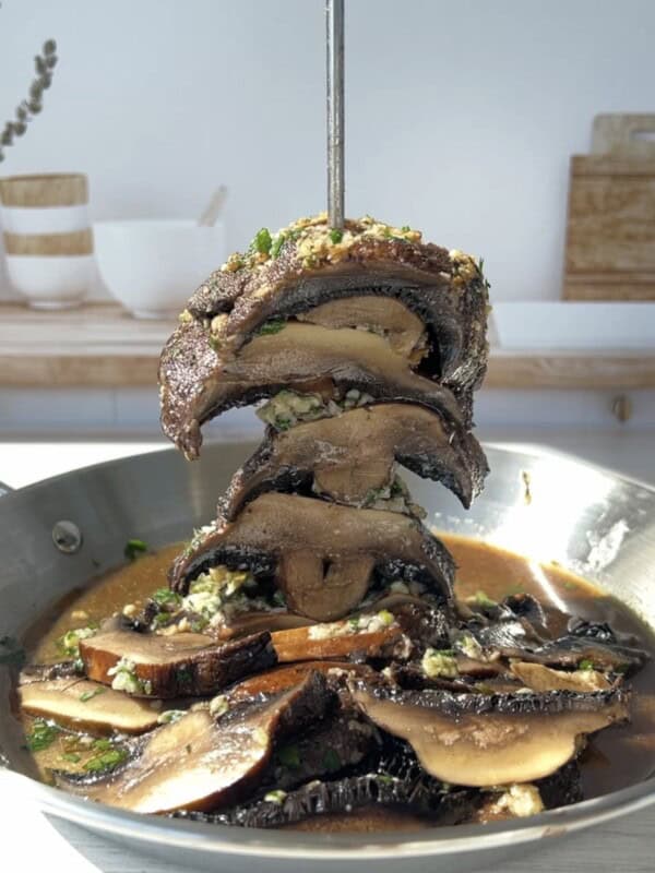 A stack of cooked portobello mushroom slices skewered vertically in a pan with garlic herb sauce, with more mushrooms and sauce at the bottom. Bright kitchen setting in the background.