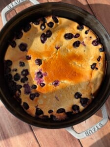 A High Protein Dutch Baby, golden-brown and baked with blueberries, is presented in a round black Staub baking dish on a wooden surface, sunlight casting soft shadows across the scene.