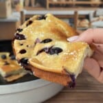A hand holds a slice of freshly baked blueberry cake, dusted with powdered sugar, while a High Protein Dutch Baby and more bread appear in the blurry background.