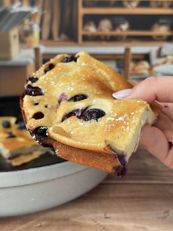 A hand holds a slice of freshly baked blueberry cake, dusted with powdered sugar, while a High Protein Dutch Baby and more bread appear in the blurry background.